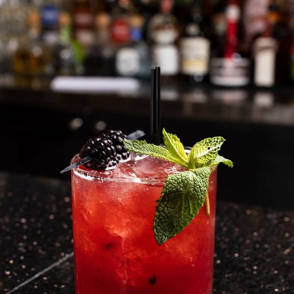 Red cocktail with blackberries, mint, and straws on a dark bar with blurred bottles in the background.