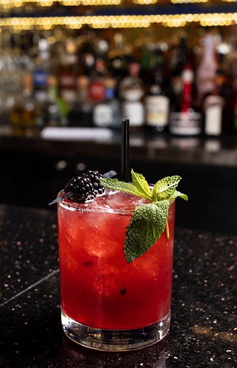 Red cocktail with blackberries, mint, and straws on a dark bar with blurred bottles in the background.