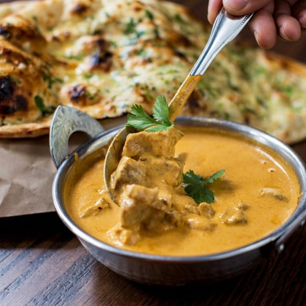 Spoon lifting chicken tikka masala from a bowl, with naan bread in the background.