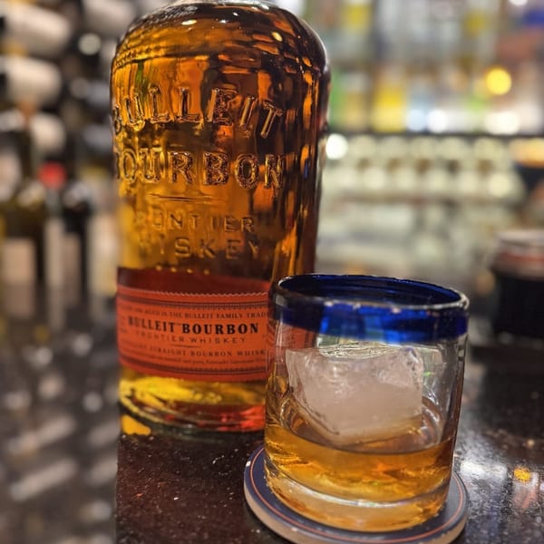 Bulleit Bourbon bottle and a glass of whiskey with ice on a dark bar top.
