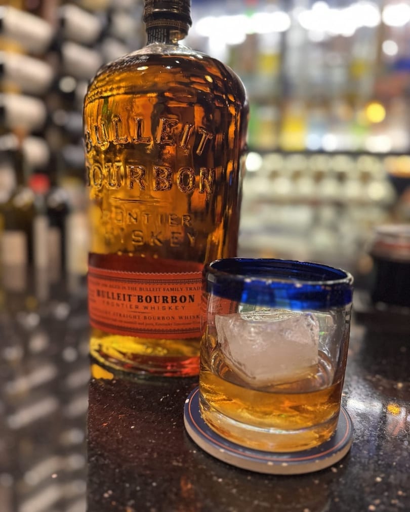 Bulleit Bourbon bottle and a glass of whiskey with ice on a dark bar top.