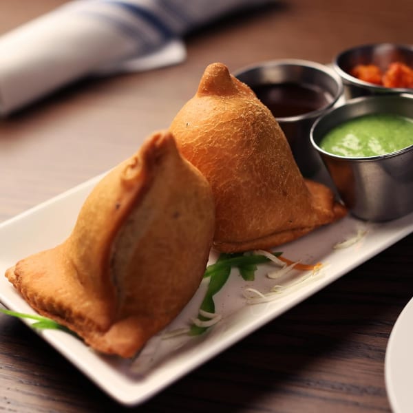 Two golden-brown samosas on a white plate with green and brown dipping sauces in steel cups.