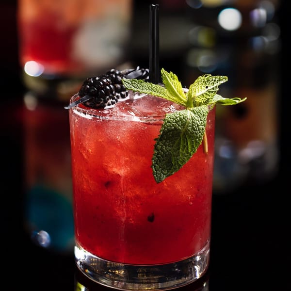 A vibrant red cocktail with ice, garnished with fresh mint and blackberries, against a dark background.