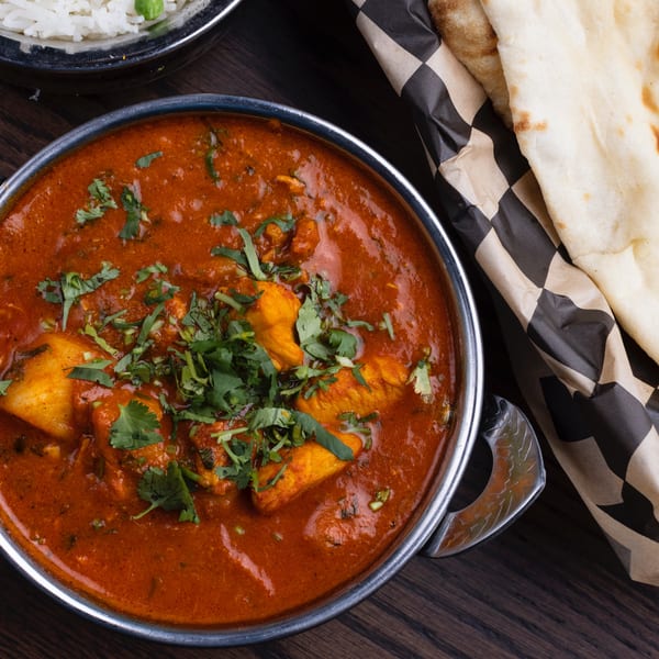 Chicken curry in a metal bowl with rice and naan bread on a wooden table.