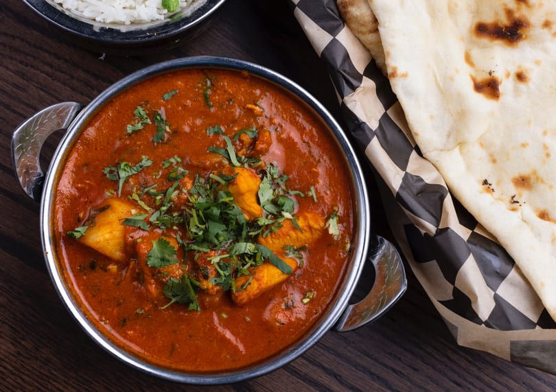 Chicken curry in a metal bowl with rice and naan bread on a wooden table.