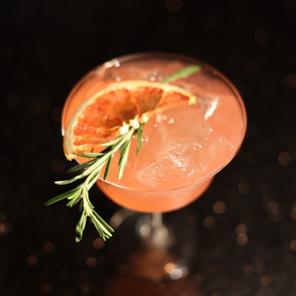 Pink cocktail with ice, garnished with a dried orange slice and a sprig of rosemary.